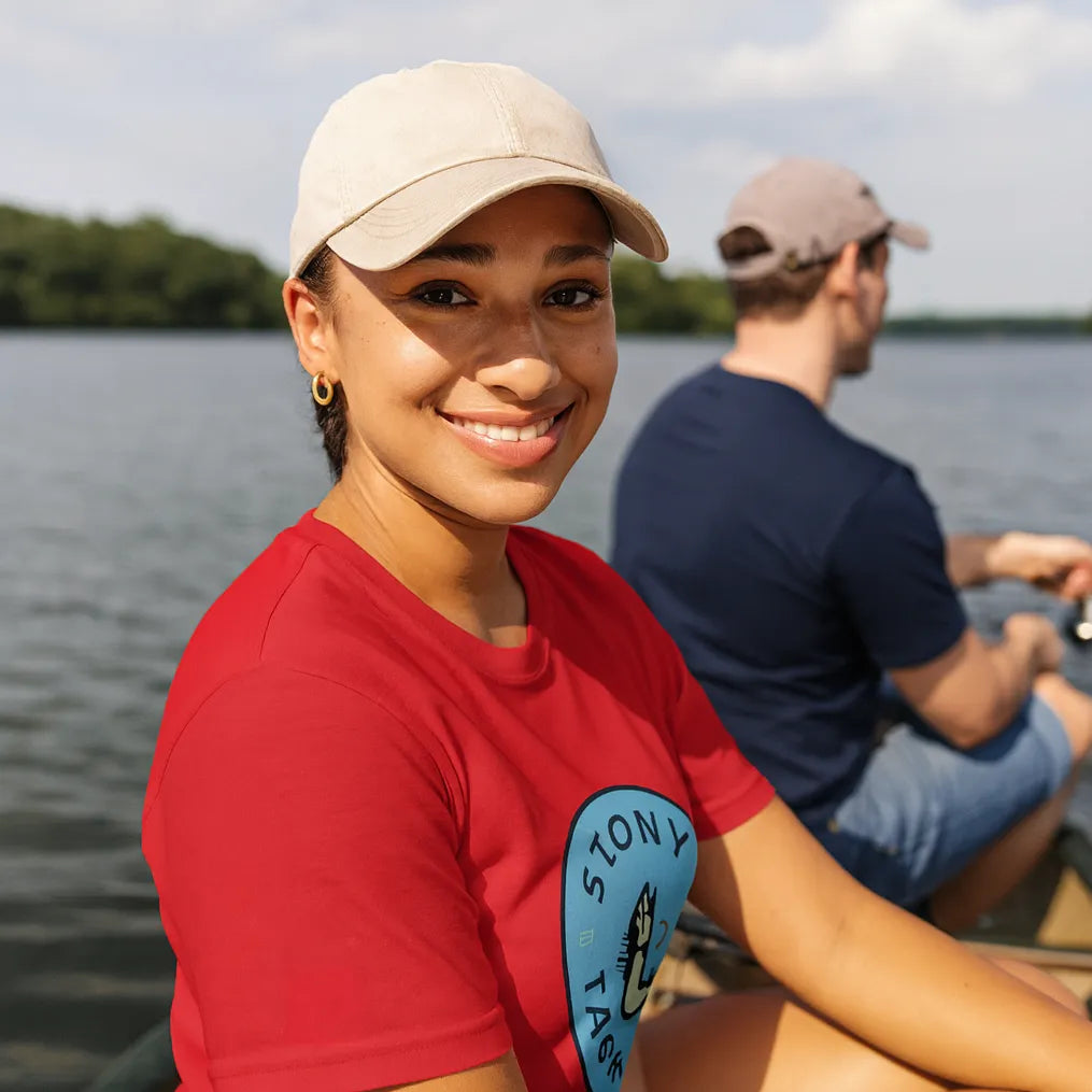 Person wearing a red shirt with a logo, sitting by a lake with another person in the background.