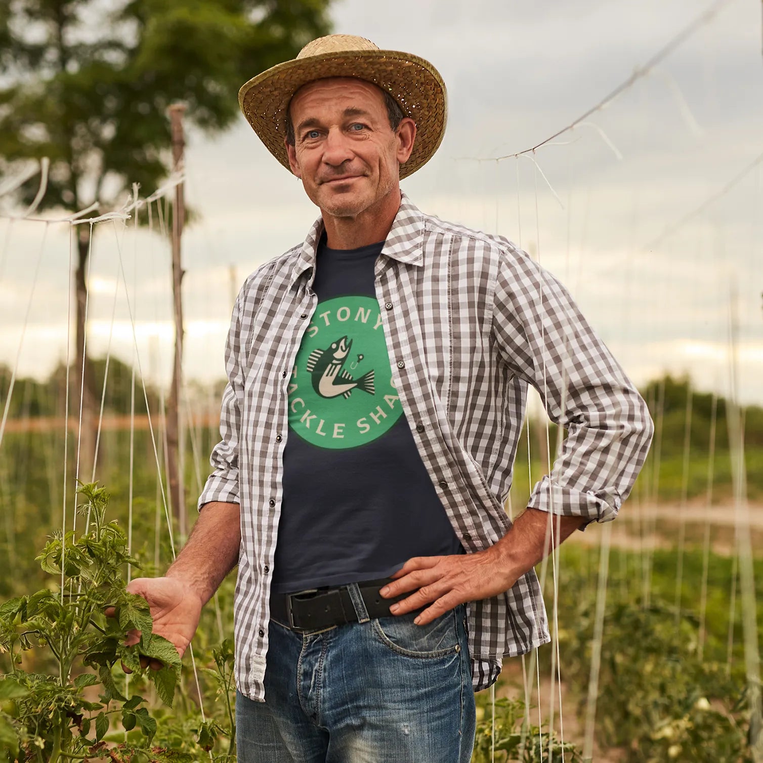 Man standing in a field with plants, wearing a hat and plaid outer shirt over a blue t shirt with a green logo