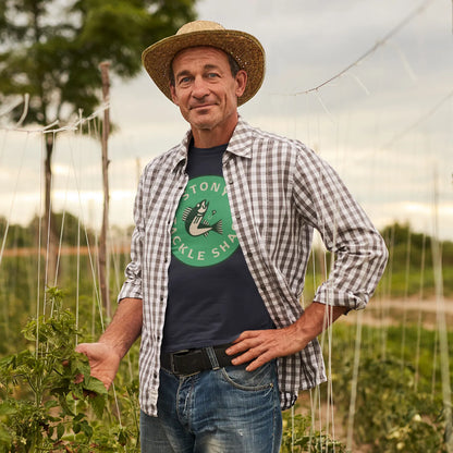 Man standing in a field with plants, wearing a hat and plaid outer shirt over a blue t shirt with a green logo