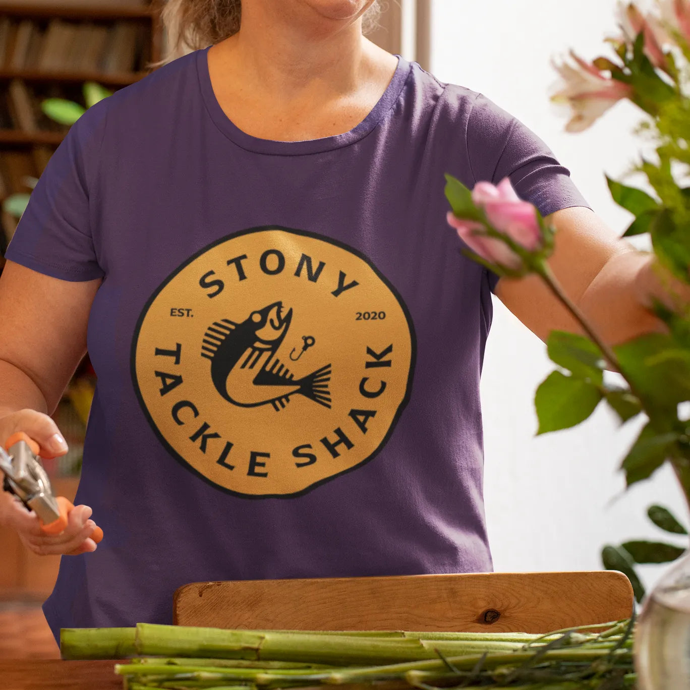 Person wearing a purple t-shirt with yellow'Stony Tackle Shack' logo, arranging flowers indoors.