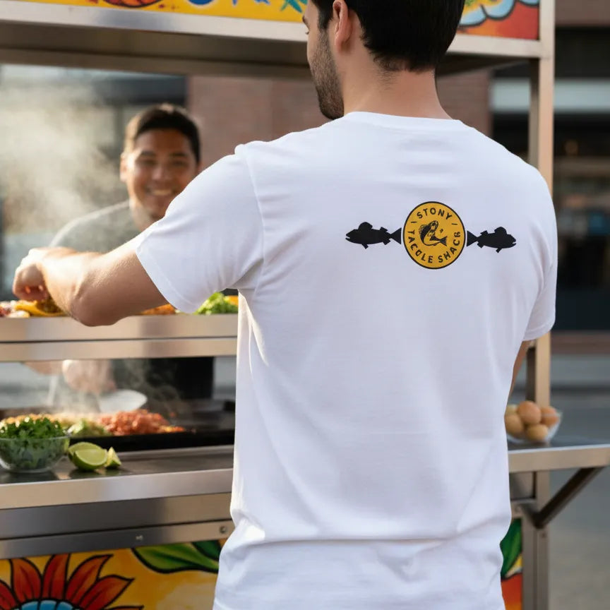Person wearing a white t-shirt with a yellow logo, buying food from a taco stand