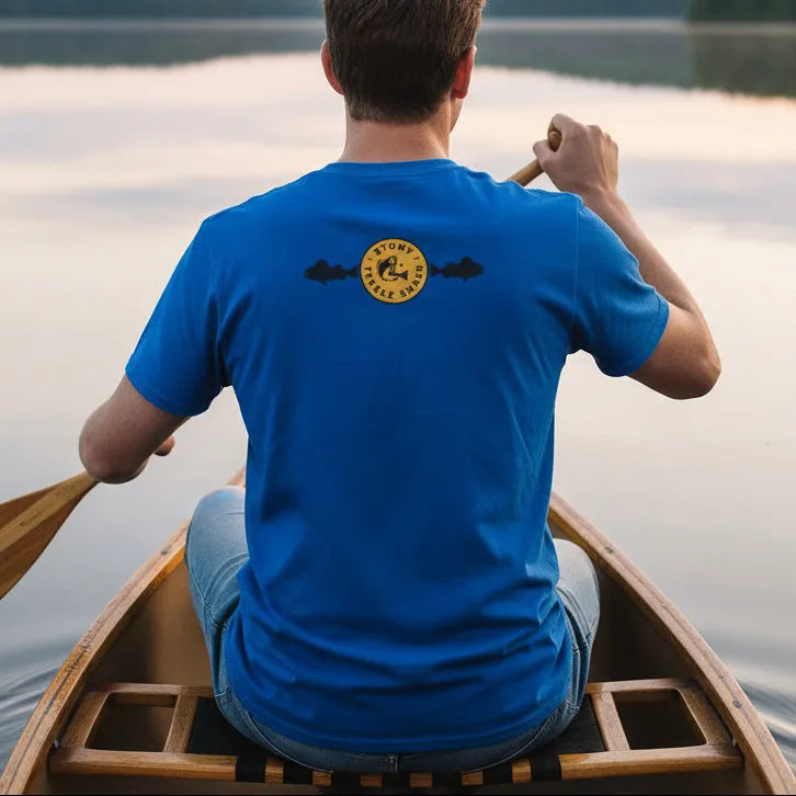 Person paddling a canoe on a lake wearing a blue t-shirt with a yellow logo.