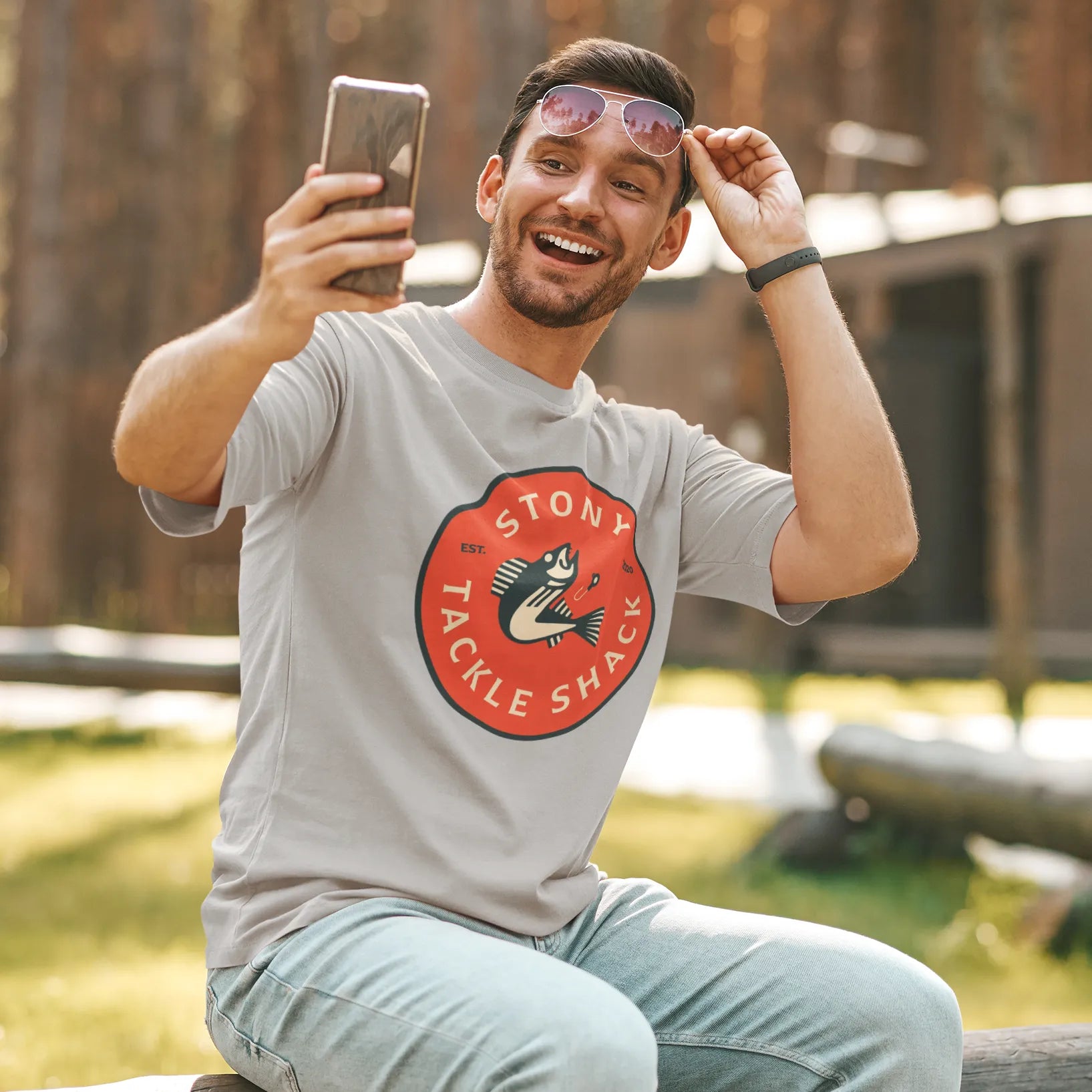 Man taking a selfie outdoors in a forested area, wearing a gray t-shirt with a red logo.
