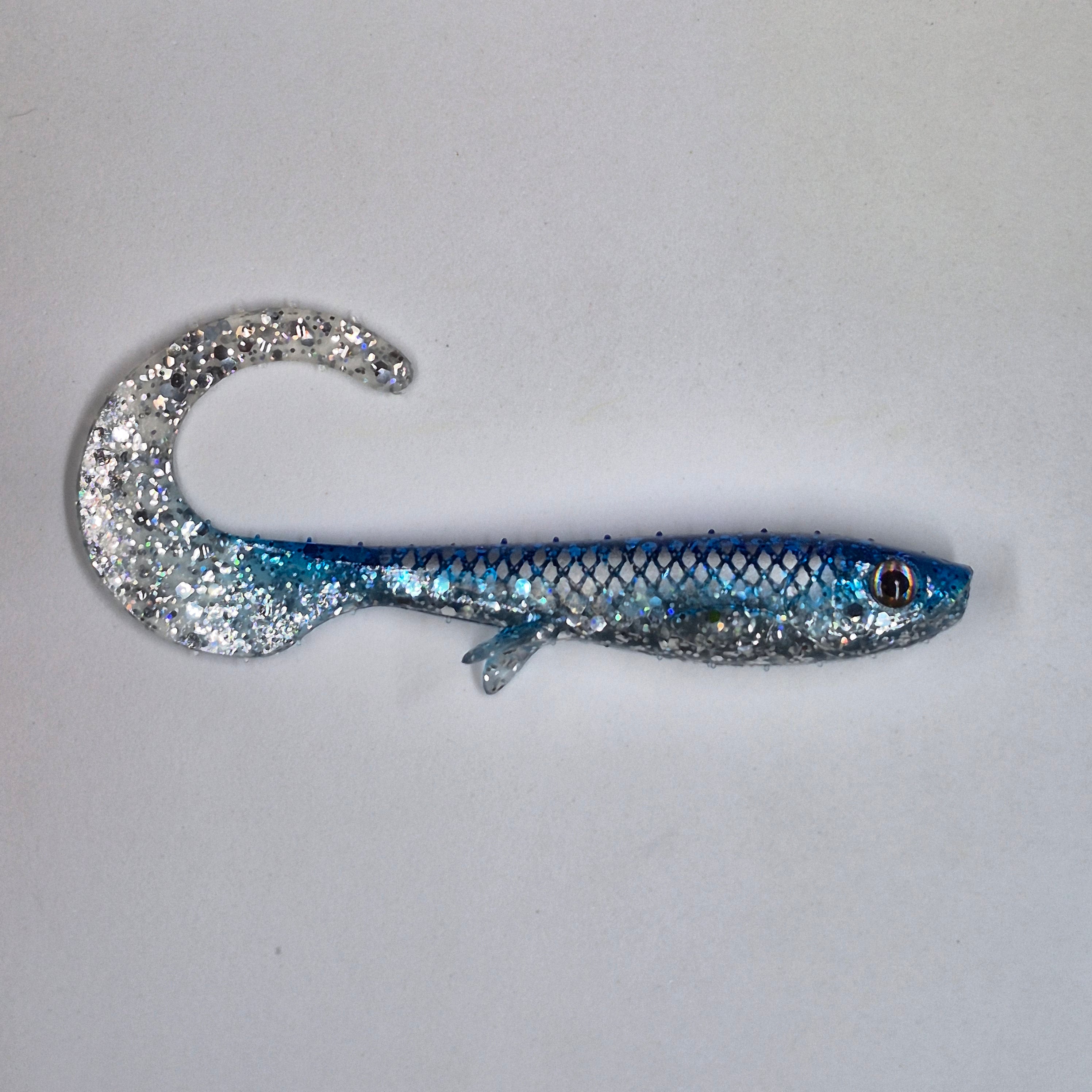 A glittery soft plastic fishing lure shaped like a small fish, featuring a curved tail with a silver glitter finish and a body that transitions from blue to silver glitter, shown on a plain light gray background.