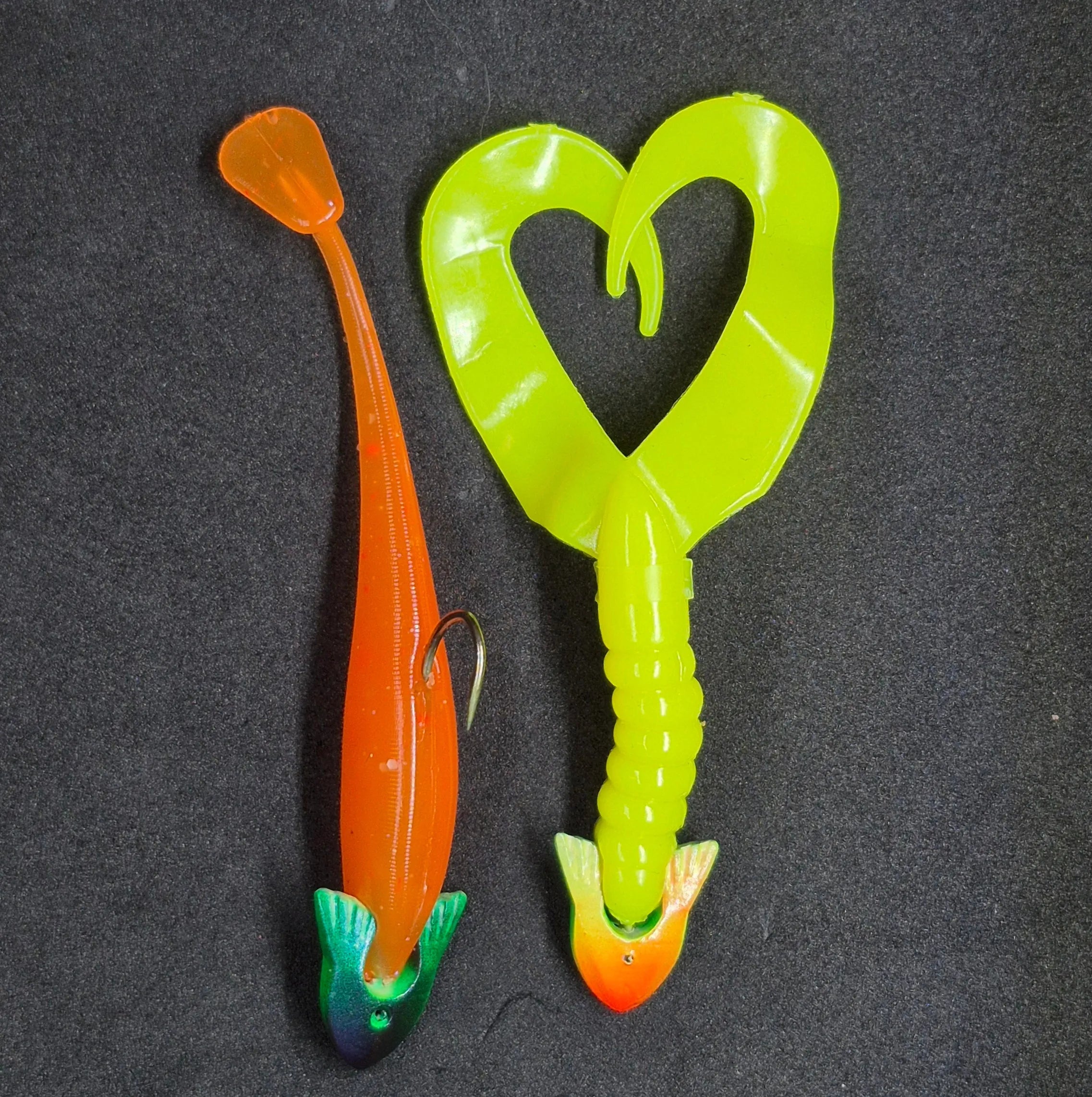 Two fishing jig heads, one orange and one green with a fin shape, on a dark background.