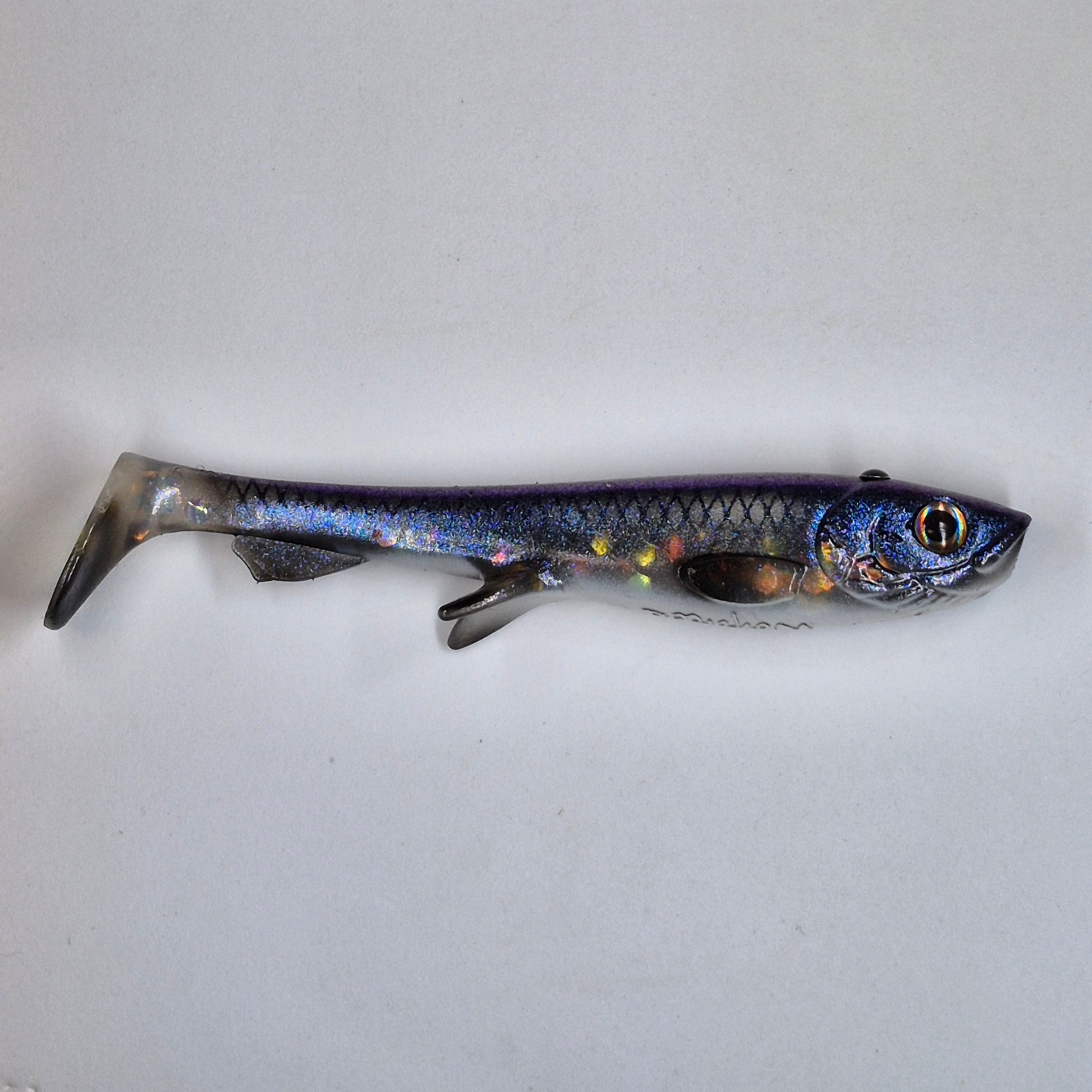 A soft plastic fishing lure shaped like a small fish, featuring a metallic blue and purple body with iridescent scales, a detailed head with a realistic eye, and a forked tail, shown against a plain light gray background.