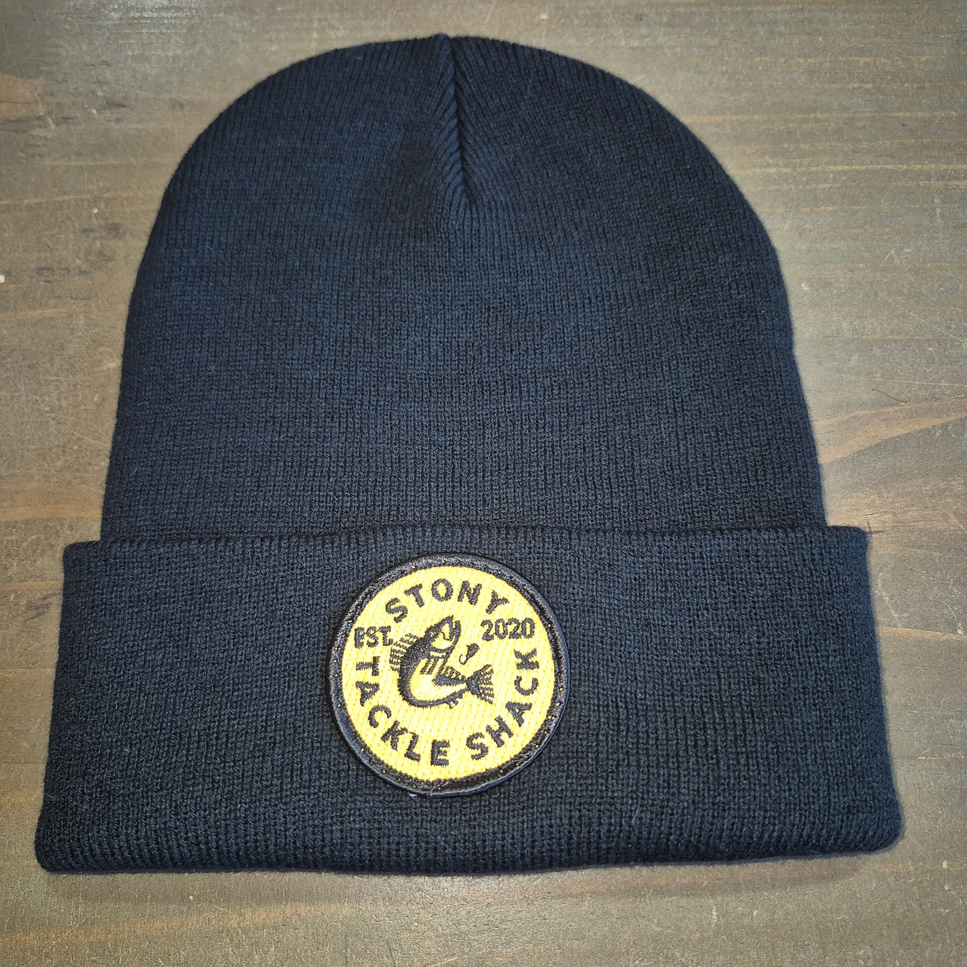 Black knit beanie with a yellow logo patch on a wooden surface