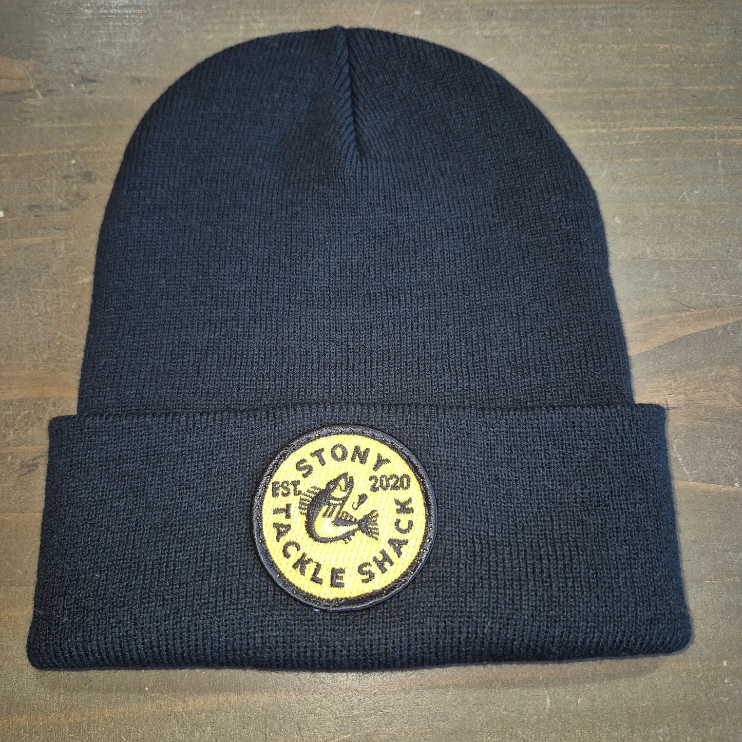 Black knit beanie with a yellow logo patch on a wooden surface