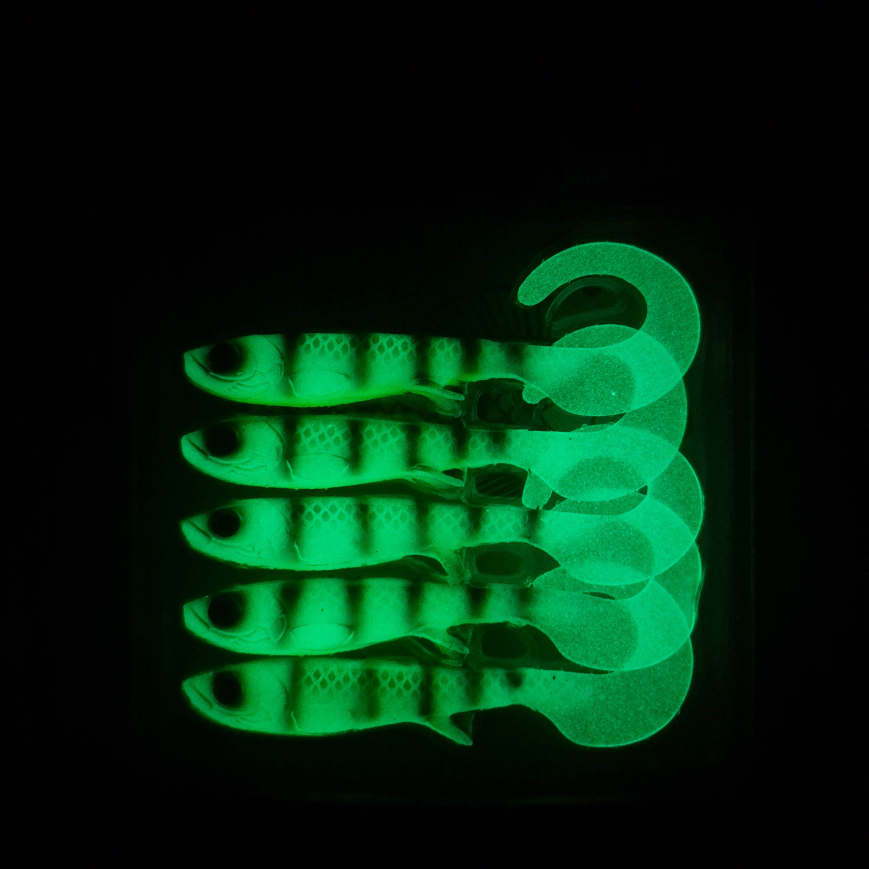 Five glowing green fishing lures with a fish design with curly tails arranged in a stack against a solid black background.