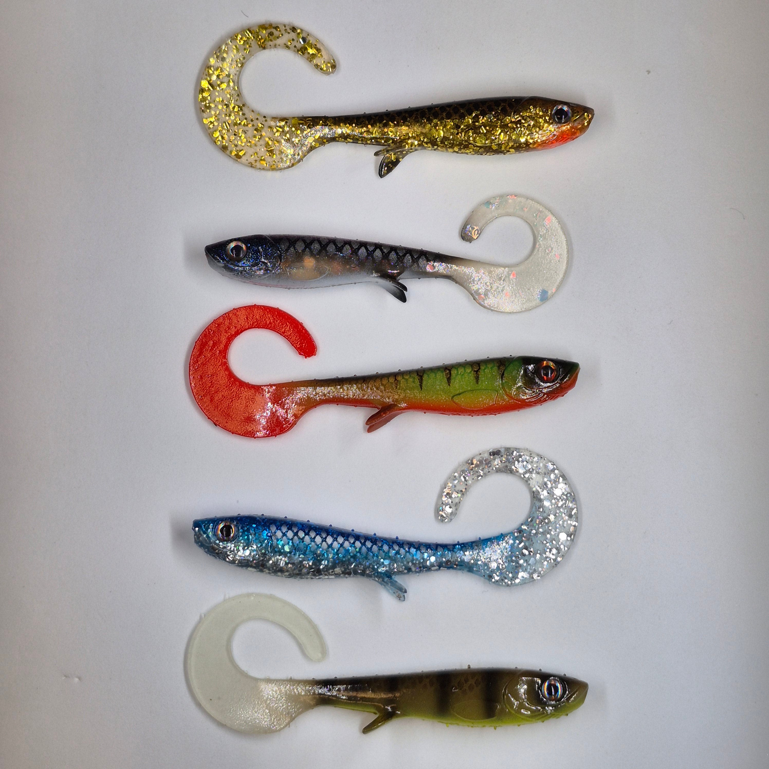 Five soft plastic fishing lures shaped like small fish with curved tails, arranged vertically on a plain white background. The lures show different color combinations and finishes, including gold glitter, blue and silver, red and green, blue and silver, and olive green and black.
