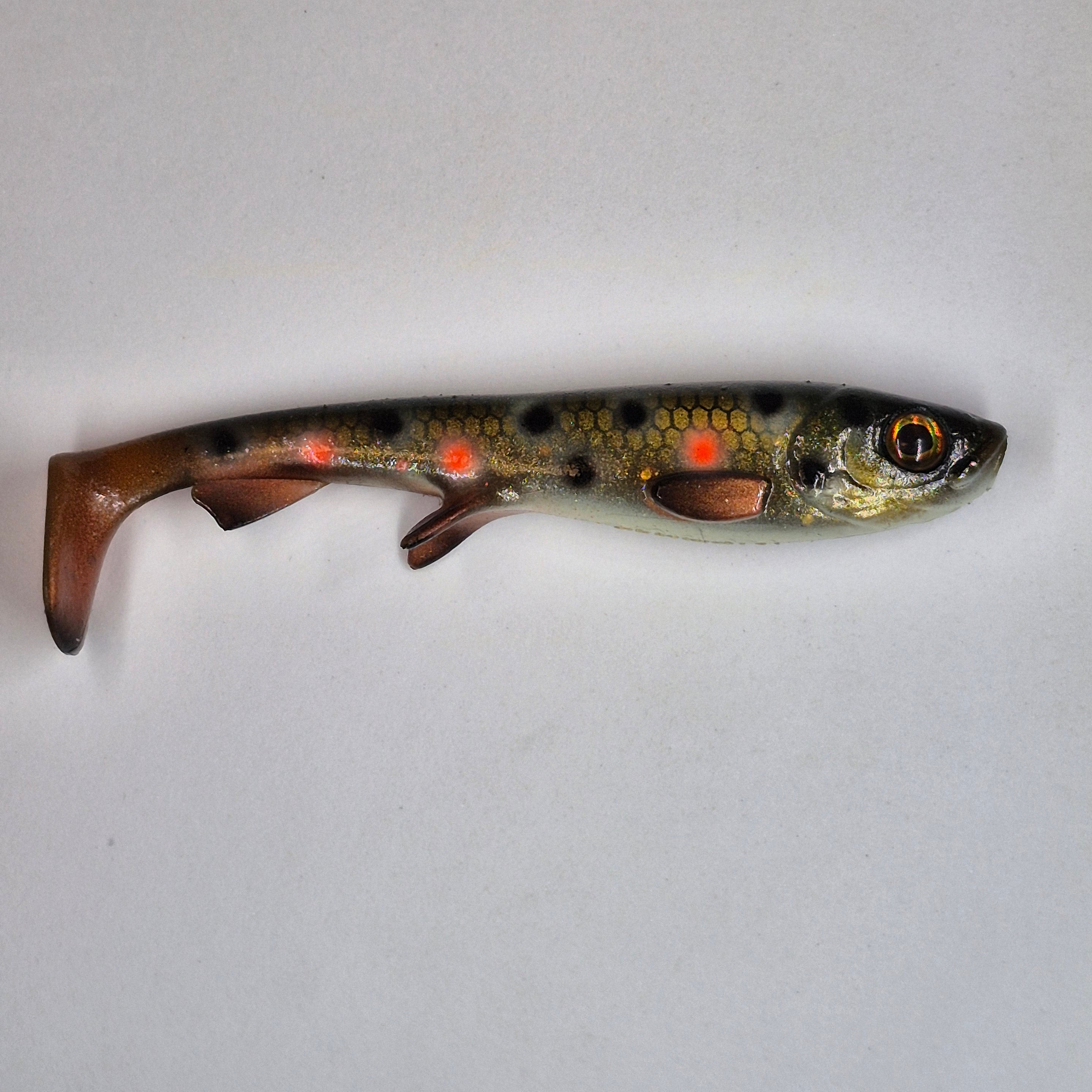 A soft plastic fishing lure shaped like a small trout, featuring a realistic scale pattern in olive green, black, and copper-brown, with bright orange-red reflective dots along its side, photographed on a plain light gray background.
