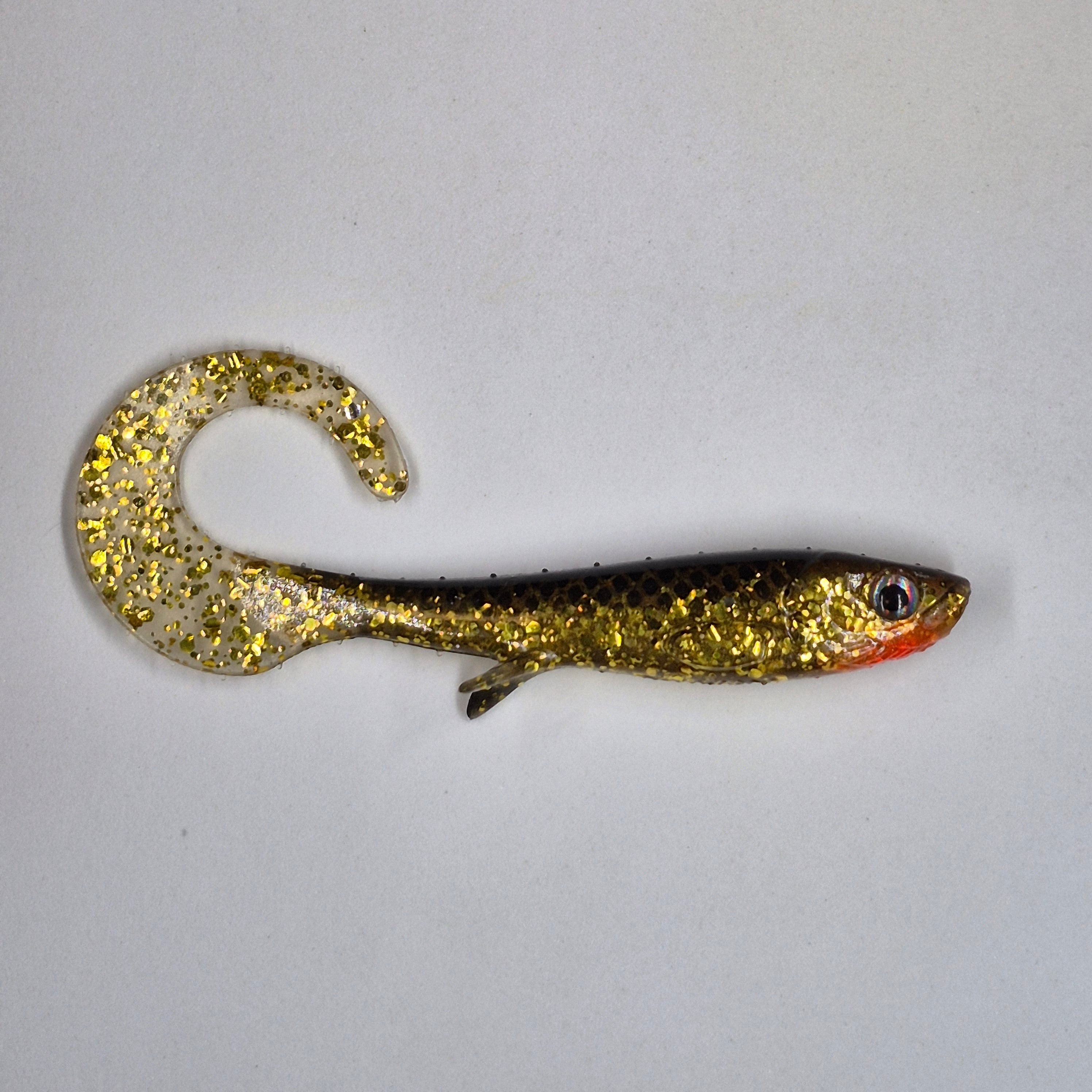 A soft plastic fishing lure shaped like a small fish, featuring a glittery gold and black body with a translucent curved tail and a red-orange mouth, shown on a plain light background.