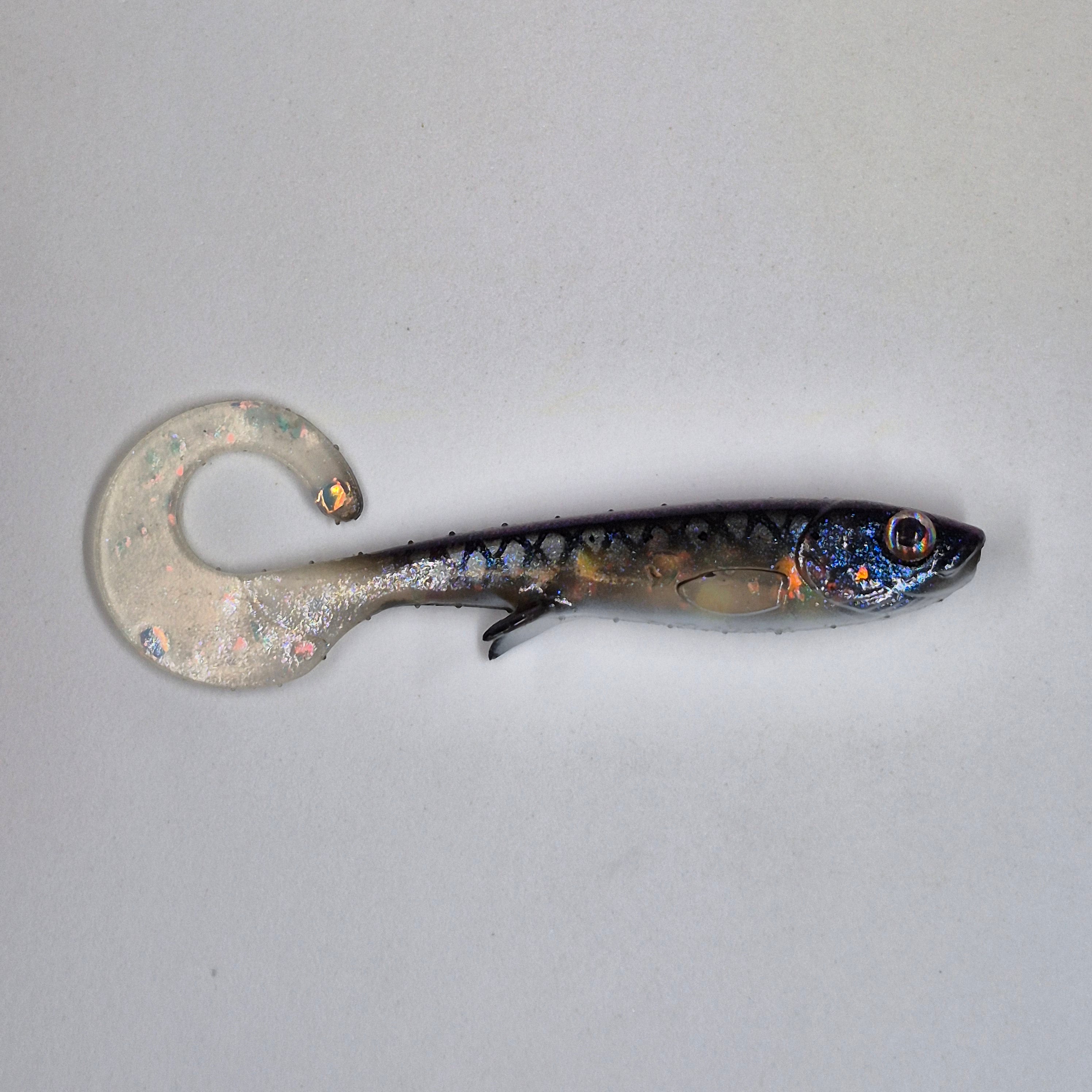 A soft plastic fishing lure shaped like a small fish with a curved tail, shown on a plain light gray background. The lure has a translucent clear body, a dark blue and black scaled back, and iridescent glitter details.